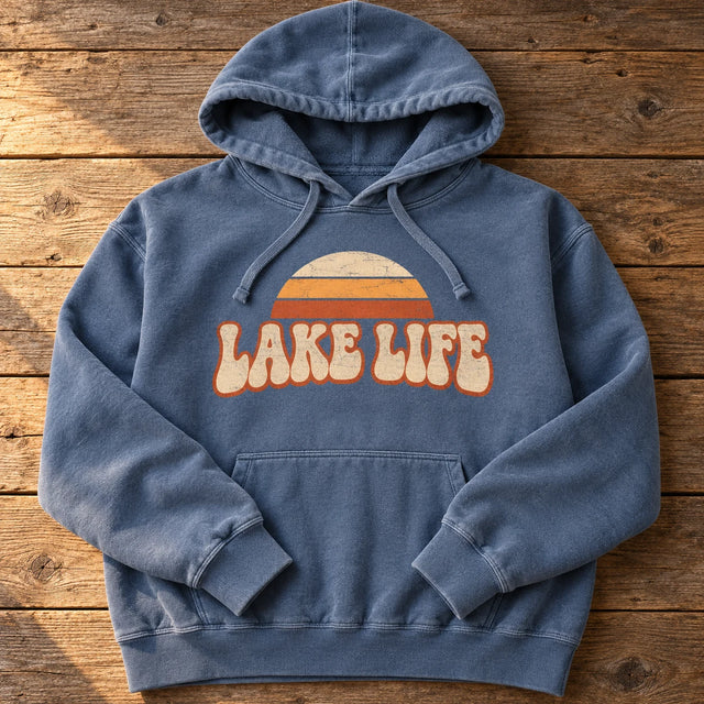 Custom printed Comfort Colors 1567 garment-dyed hoodie in Blue Jean with a retro LAKE LIFE sunset design on the chest, laid flat on weathered wood