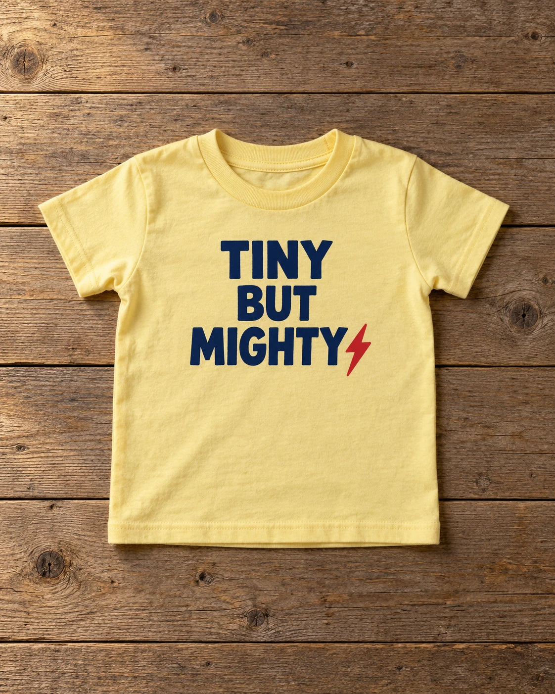 Custom butter yellow Rabbit Skins toddler t-shirt printed with a navy Tiny But Mighty design, laid flat on weathered wood
