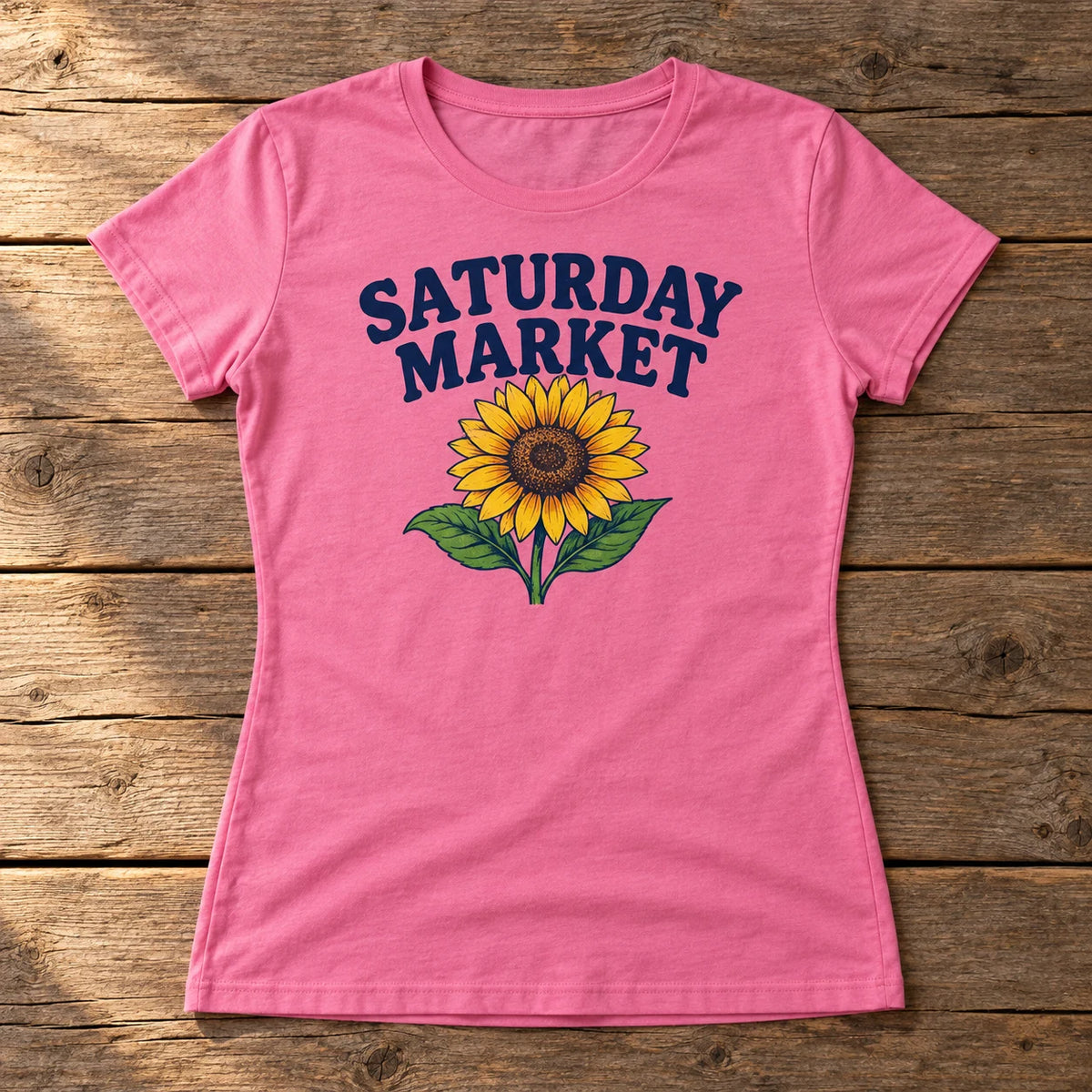 Custom women's Softstyle t-shirt in azalea pink with a full color Saturday Market sunflower DTF print, on a wood table