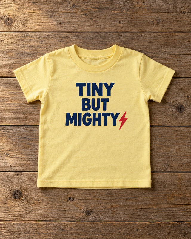 Custom butter yellow Rabbit Skins toddler t-shirt printed with a navy Tiny But Mighty design, laid flat on weathered wood