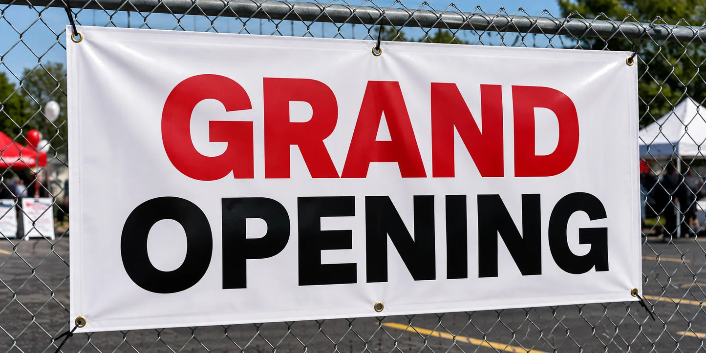 Custom 13 oz vinyl banner with hemmed edges and brass grommets reading Grand Opening, zip-tied to a chain-link fence