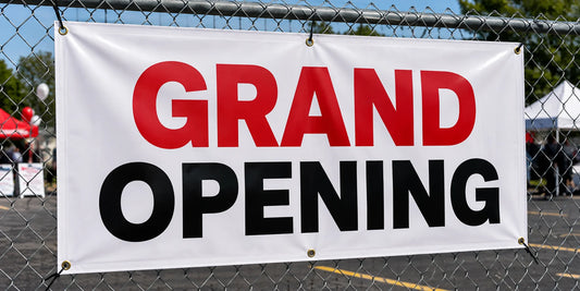 Custom 13 oz vinyl banner with hemmed edges and brass grommets reading Grand Opening, zip-tied to a chain-link fence