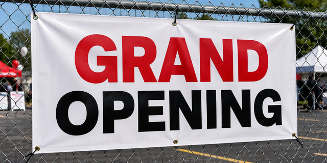 Custom 13 oz vinyl banner with hemmed edges and brass grommets reading Grand Opening, zip-tied to a chain-link fence