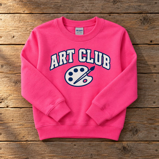 Custom Gildan Heavy Blend youth crewneck sweatshirt in Safety Pink with a full-color ART CLUB DTF print, folded on a wood table