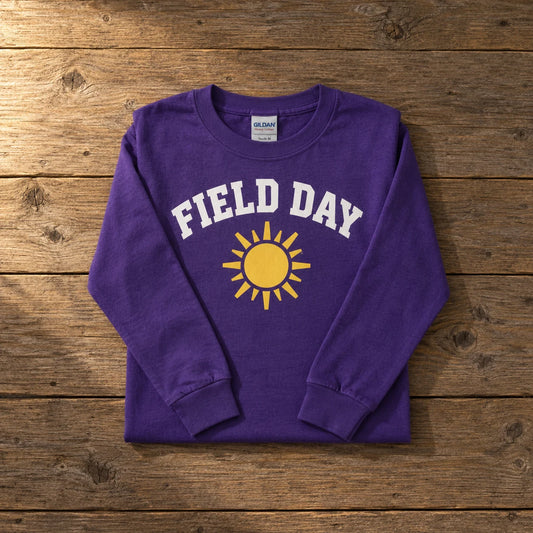 Custom Gildan Heavy Cotton youth long sleeve t-shirt in Purple with a full-color FIELD DAY sun DTF print, folded on a wood table