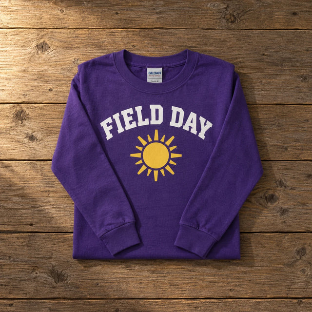 Custom Gildan Heavy Cotton youth long sleeve t-shirt in Purple with a full-color FIELD DAY sun DTF print, folded on a wood table